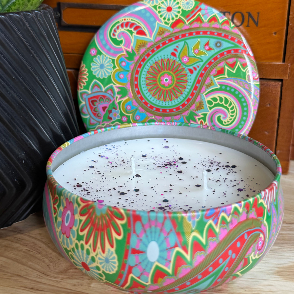 A round metal candle tin filled with white wax and topped with dark berry-like sprinkles, shown open with its lid leaning behind it. The tin and lid feature a colorful paisley and floral pattern in green, pink, orange, blue, and yellow.