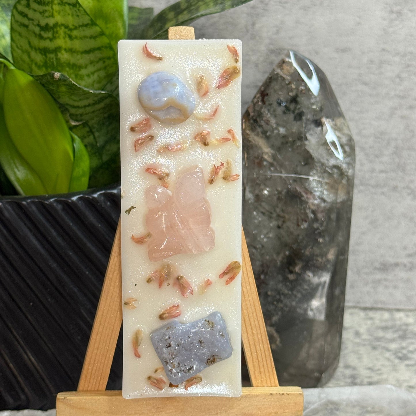 A rectangular cream-colored resin candle displayed on a small wooden easel. The candle features embedded natural stones in blue, pink, and gray, along with small dried flower buds.