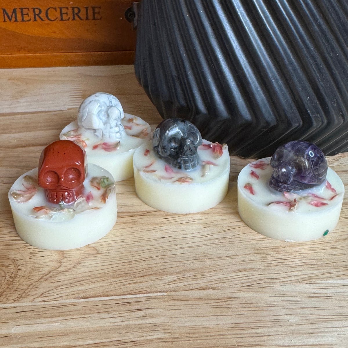 Four small oval candles with embedded resin details and skull motifs, displayed on a wooden surface in front of a black ribbed container and a wooden box labeled MERCERIE.