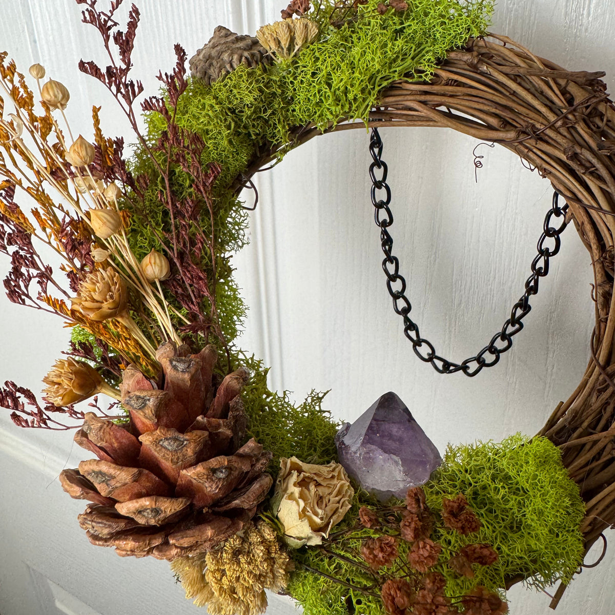 A rustic woven wicker wreath decorated with bright green moss, dried brown and tan botanicals, a pinecone, a purple crystal, and a black chain hanging on the side, shown against a white paneled door.