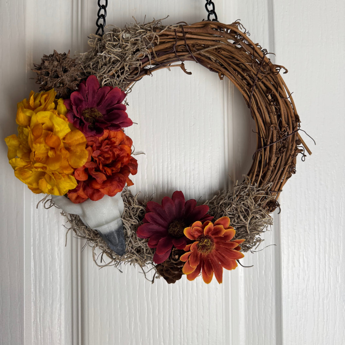 A rustic fall wreath made of woven grapevine branches, decorated with faux flowers in yellow, orange, burgundy, and red, hanging on a white paneled door.