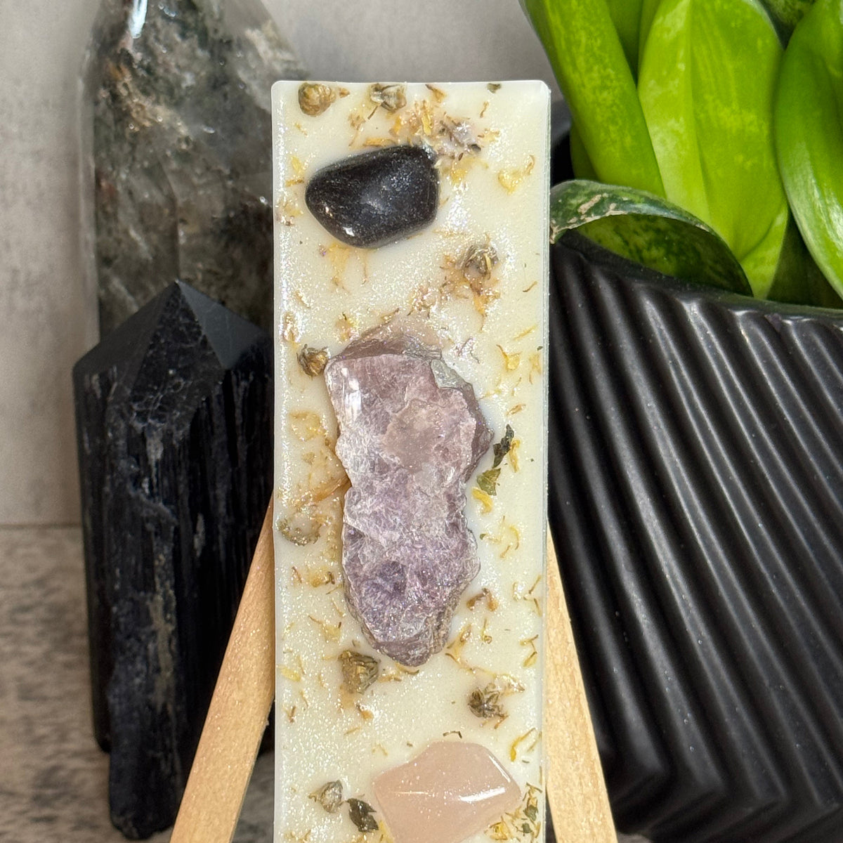 A rectangular resin candle displayed on a small wooden easel, featuring embedded natural stones and dried botanicals. The candle has a cream base with a prominent lavender-purple geode in the center, a dark black stone near the top, and a pale pink stone near the bottom.
