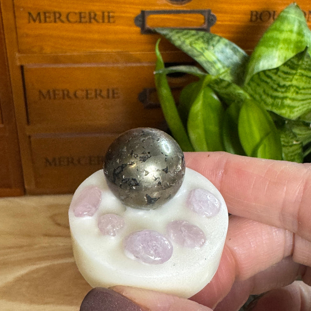 A hand holds a white circular crystal cluster with a metallic silver sphere on top, set against a wooden drawer and green plant background.