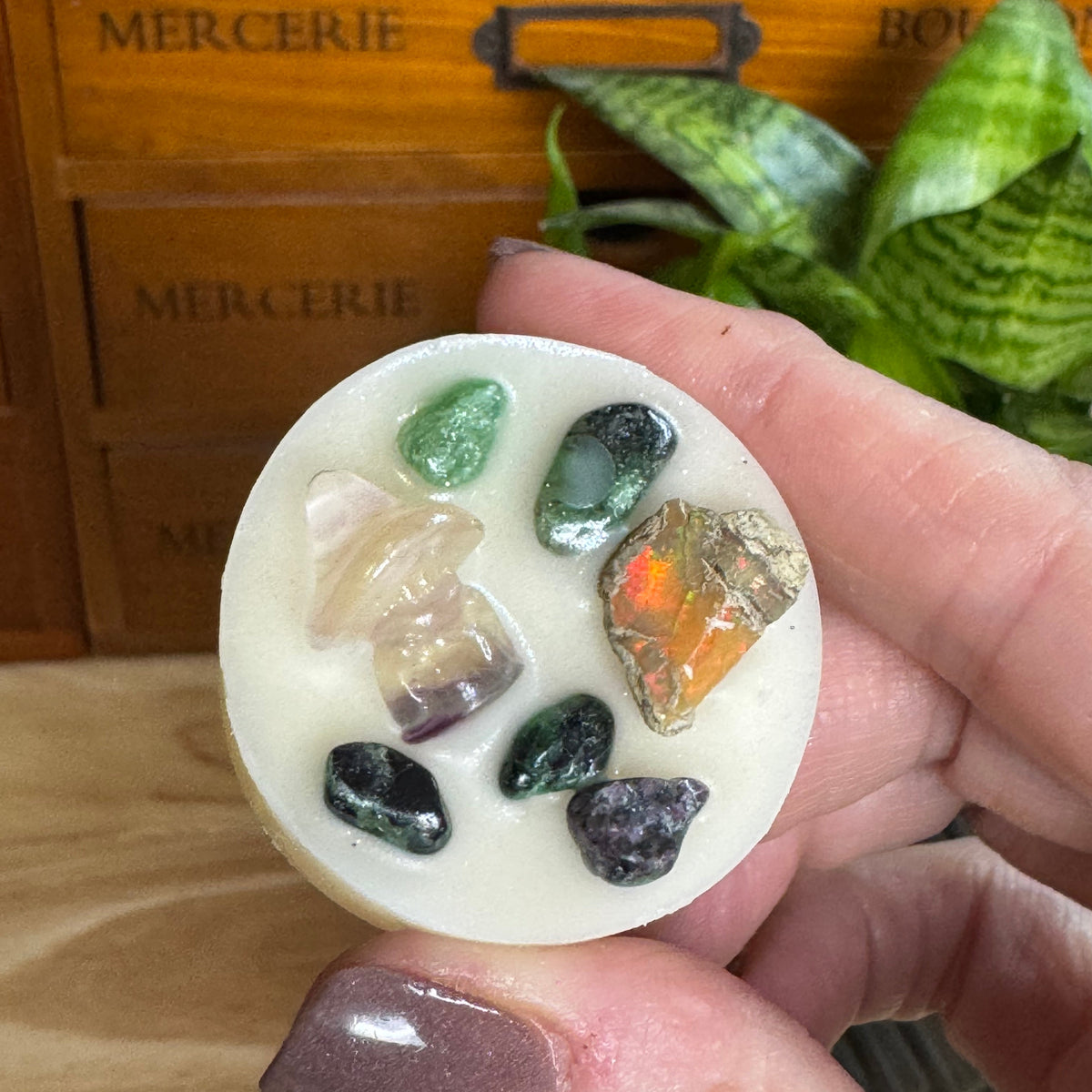 A hand with mauve nail polish holds a round white wax melt or soap insert decorated with various small tumbled and raw crystals in green, orange, purple, and clear, set against a background of a wooden drawer and a green plant.