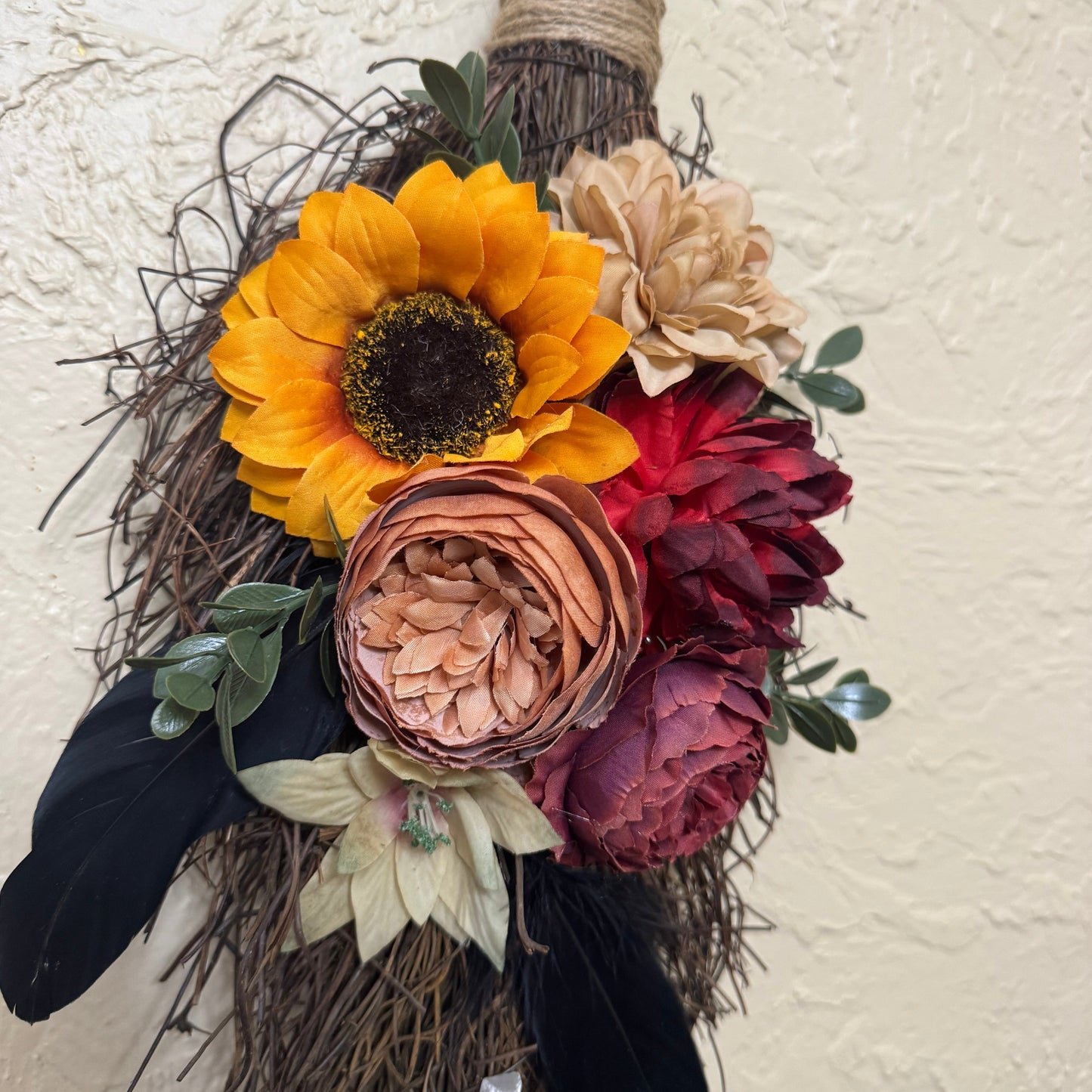 Decorative broom with autumnal flowers and feathers against a neutral wall.