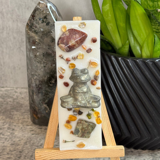 A vertical white resin candle displayed on a small wooden easel. The candle features a silver frog figure in the center, surrounded by scattered natural stones, yellow dried botanicals, and a brown polished chip near the top.