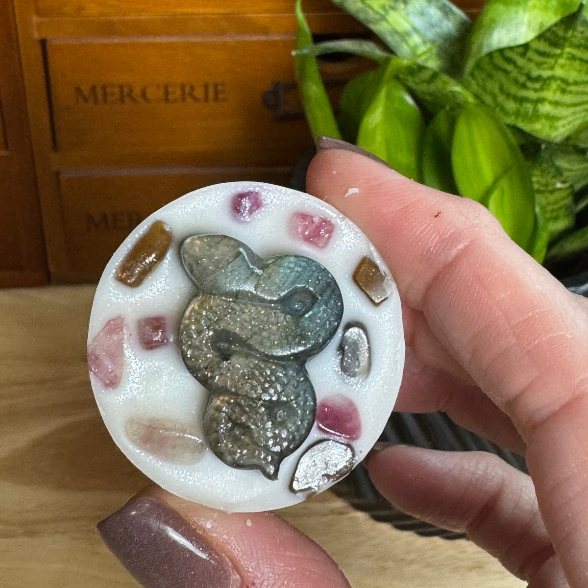 A hand holding a round white resin coaster featuring a raised metallic silver snake motif surrounded by small pink, brown, and gray decorative stones. The background shows a wooden drawer with the word 'MERCERIE' and a green plant.