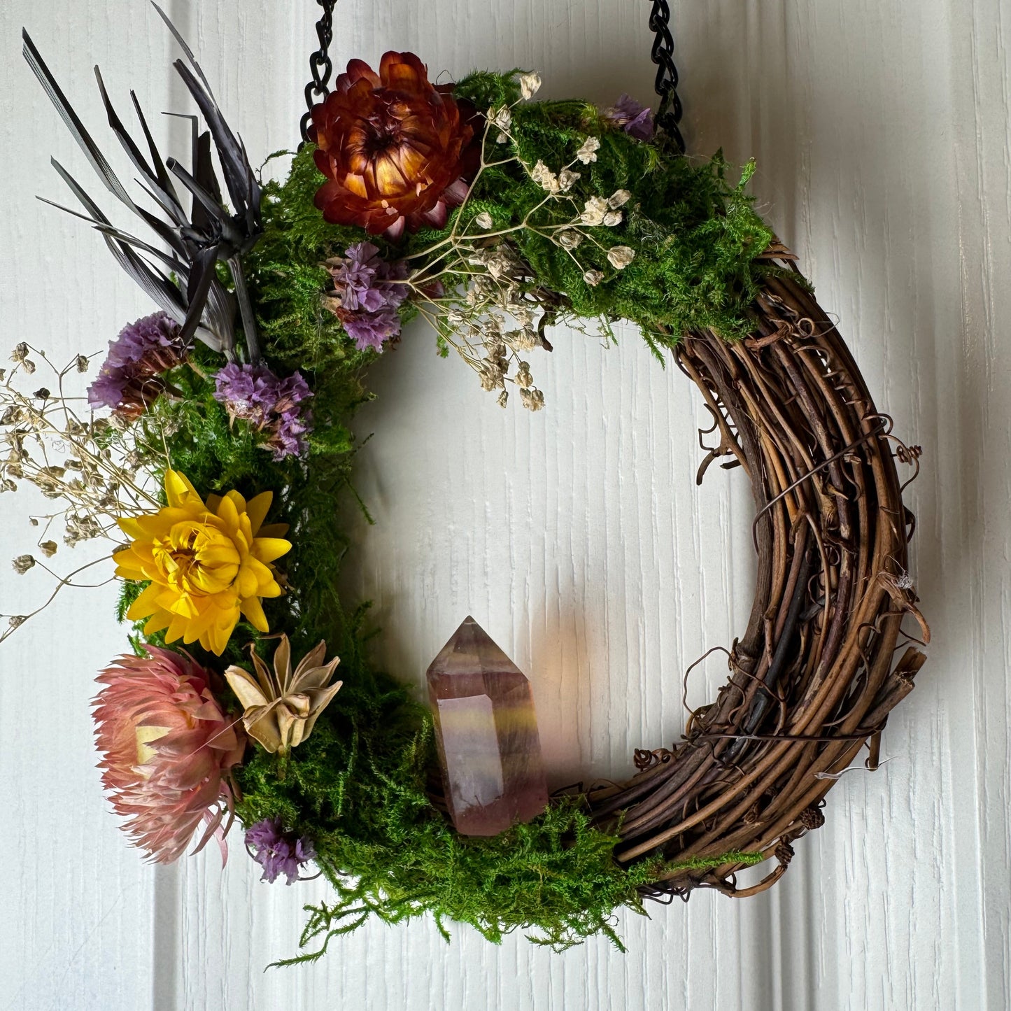 Decorative wreath with flowers, moss, and a crystal on a white door.