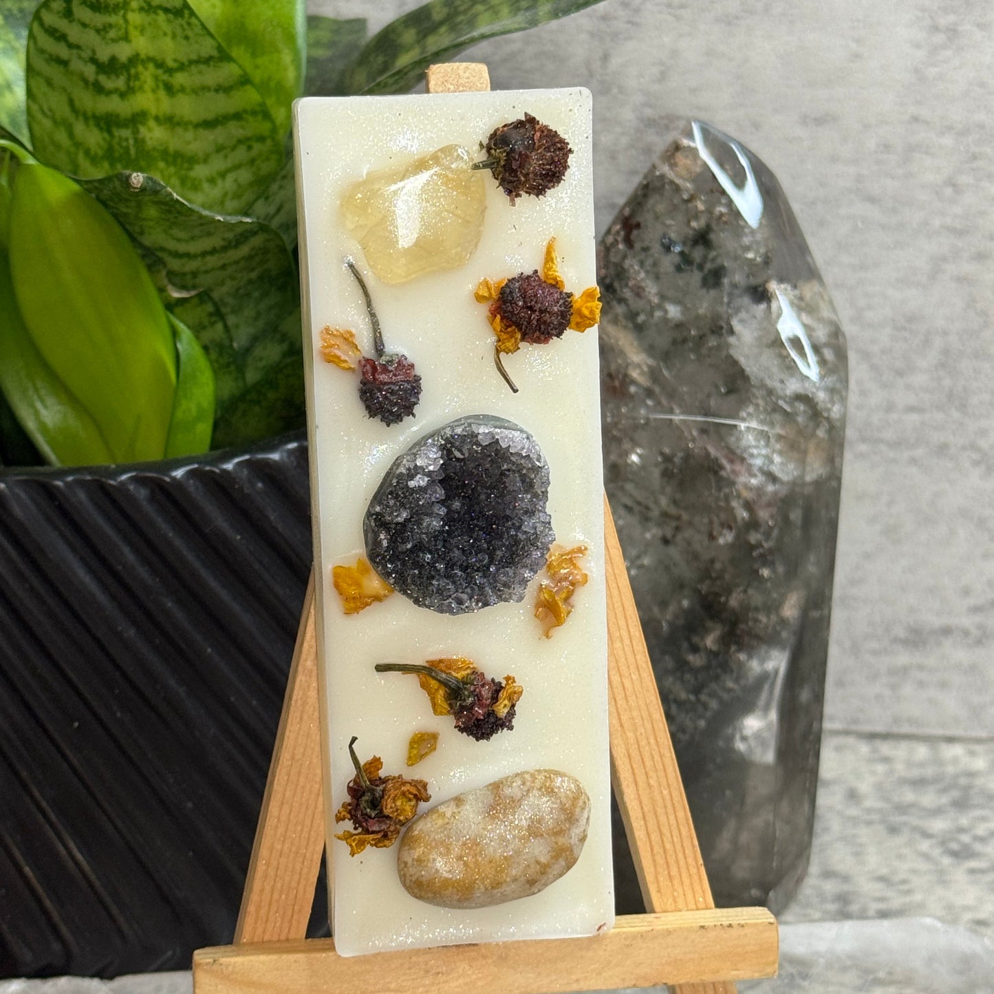 A rectangular white soap bar with embedded stones and dried flowers, displayed on a small wooden easel. The soap features a large dark gray stone, a smooth beige stone, and smaller botanical inclusions.