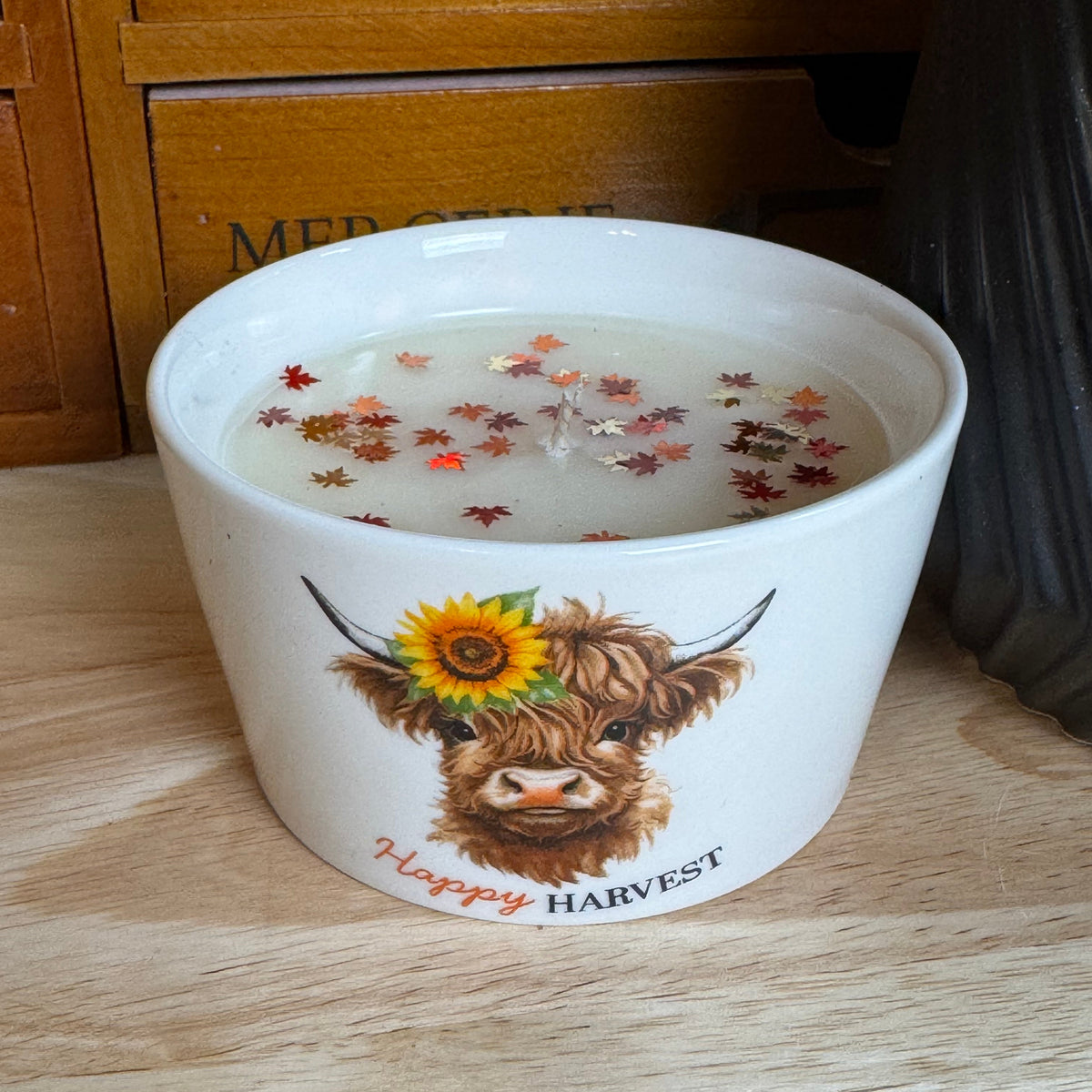 A white ceramic candle jar with a Highland cow illustration and 'Happy HARVEST' text, filled with wax and decorated with dried autumn leaves, sitting on a wooden surface.