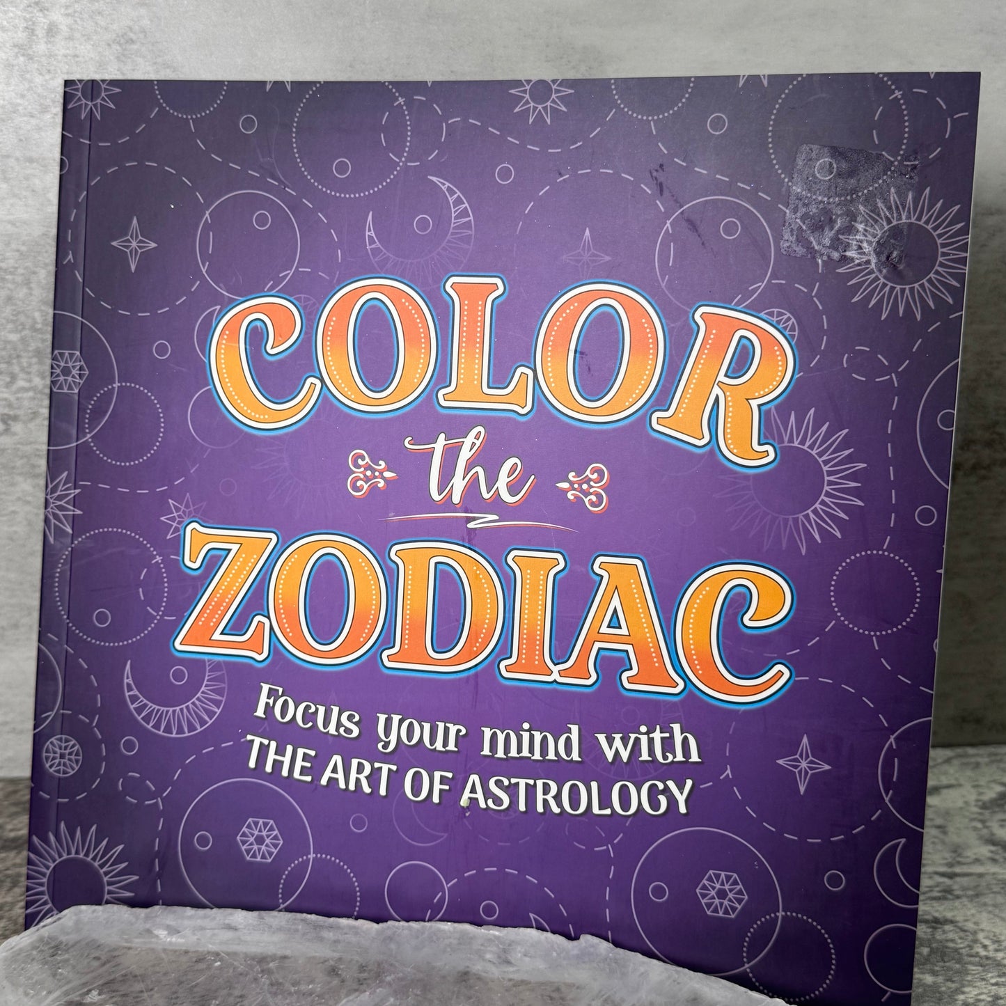 Book titled 'Color the Zodiac' on a textured surface with a stone-like object.