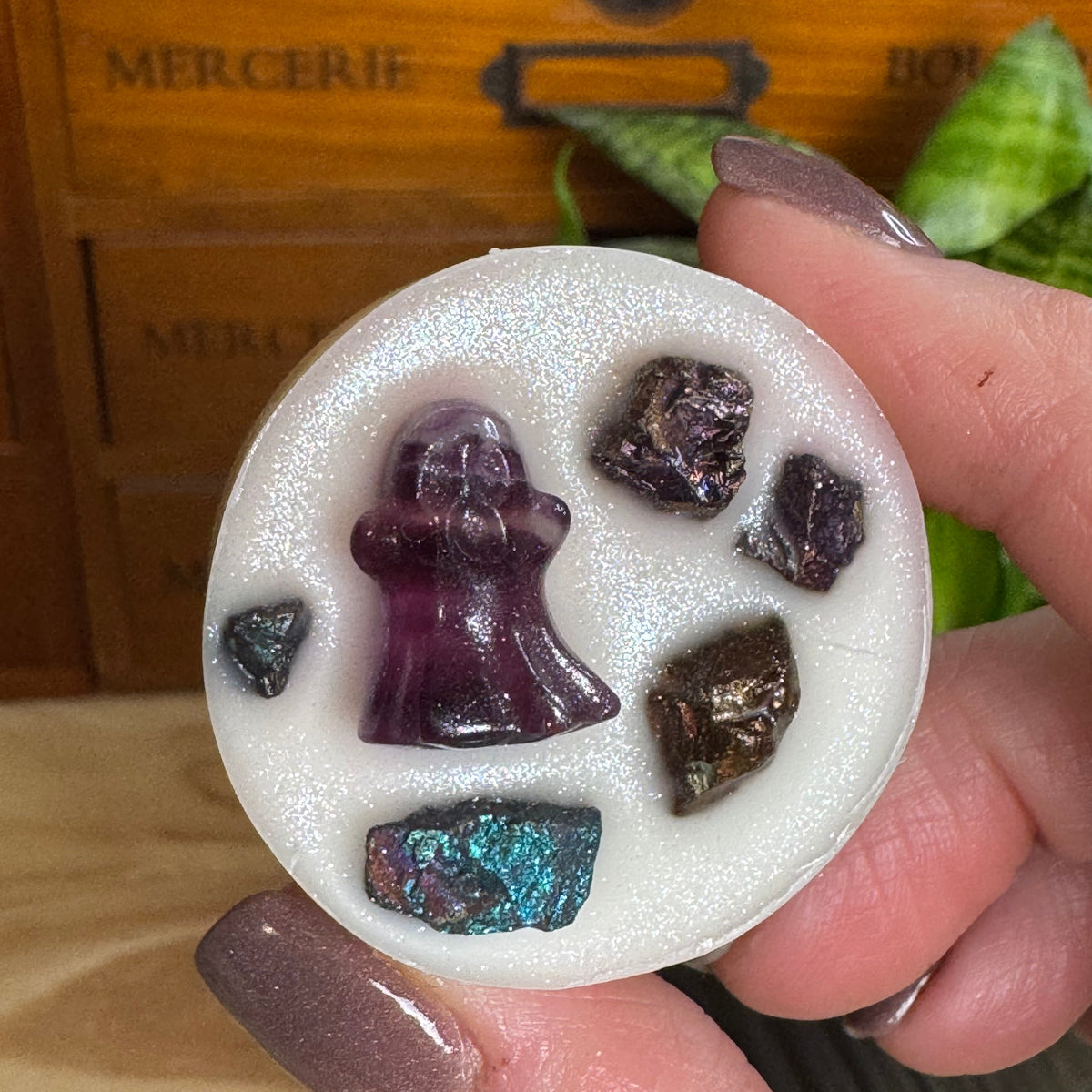 A hand with glittery mauve nails holds a round white wax melt or soap featuring a purple angel figure and several rough crystal stones, with a wooden drawer and green plant in the background.
