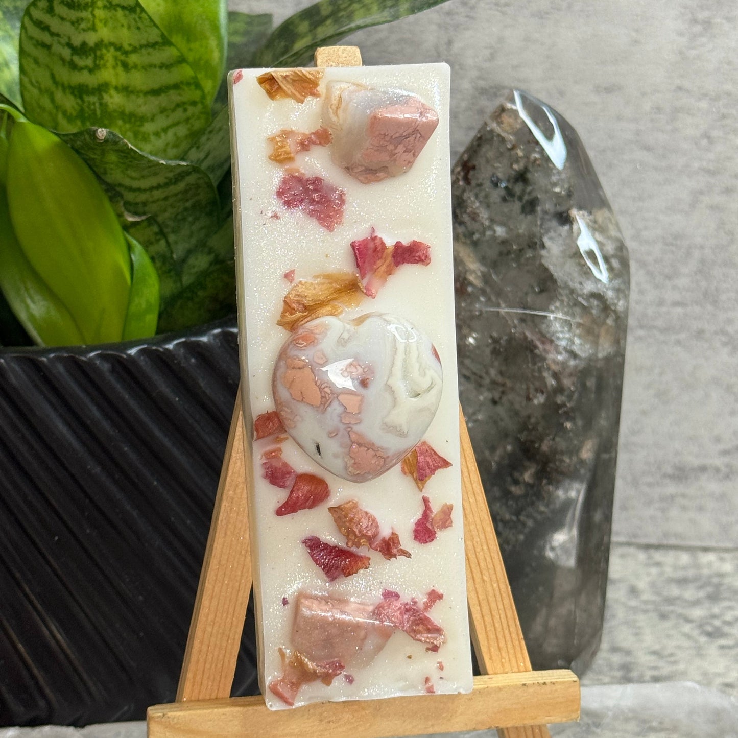 A rectangular white soap bar with embedded pink and peach stone pieces and dried flower petals, displayed on a small wooden easel. A green snake plant in a black pot and a large clear crystal are visible in the background against a gray textured wall.
