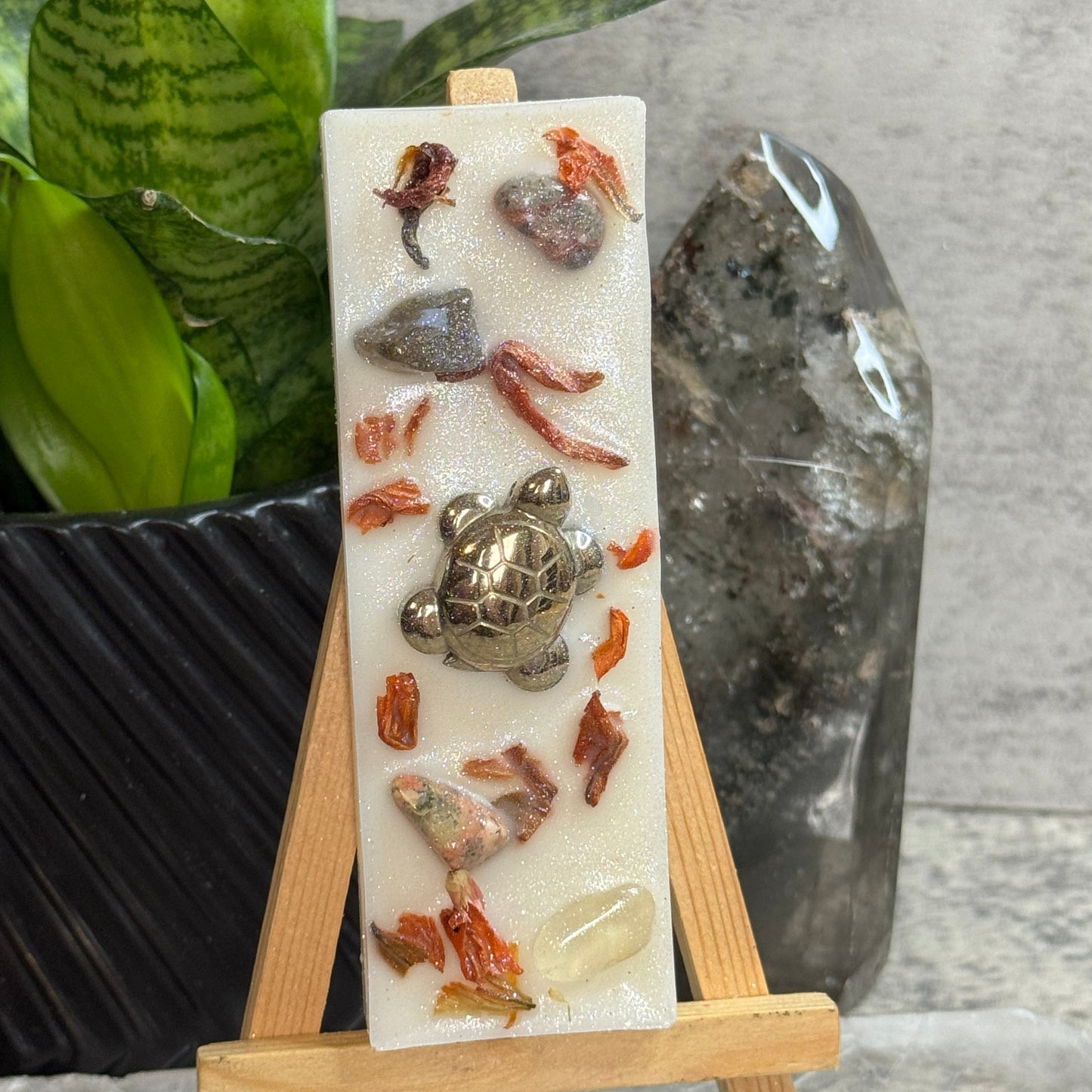 A rectangular white resin candle displayed on a small wooden easel. The candle features a raised metallic turtle figure and scattered dried botanical pieces in orange, brown, and gray tones.