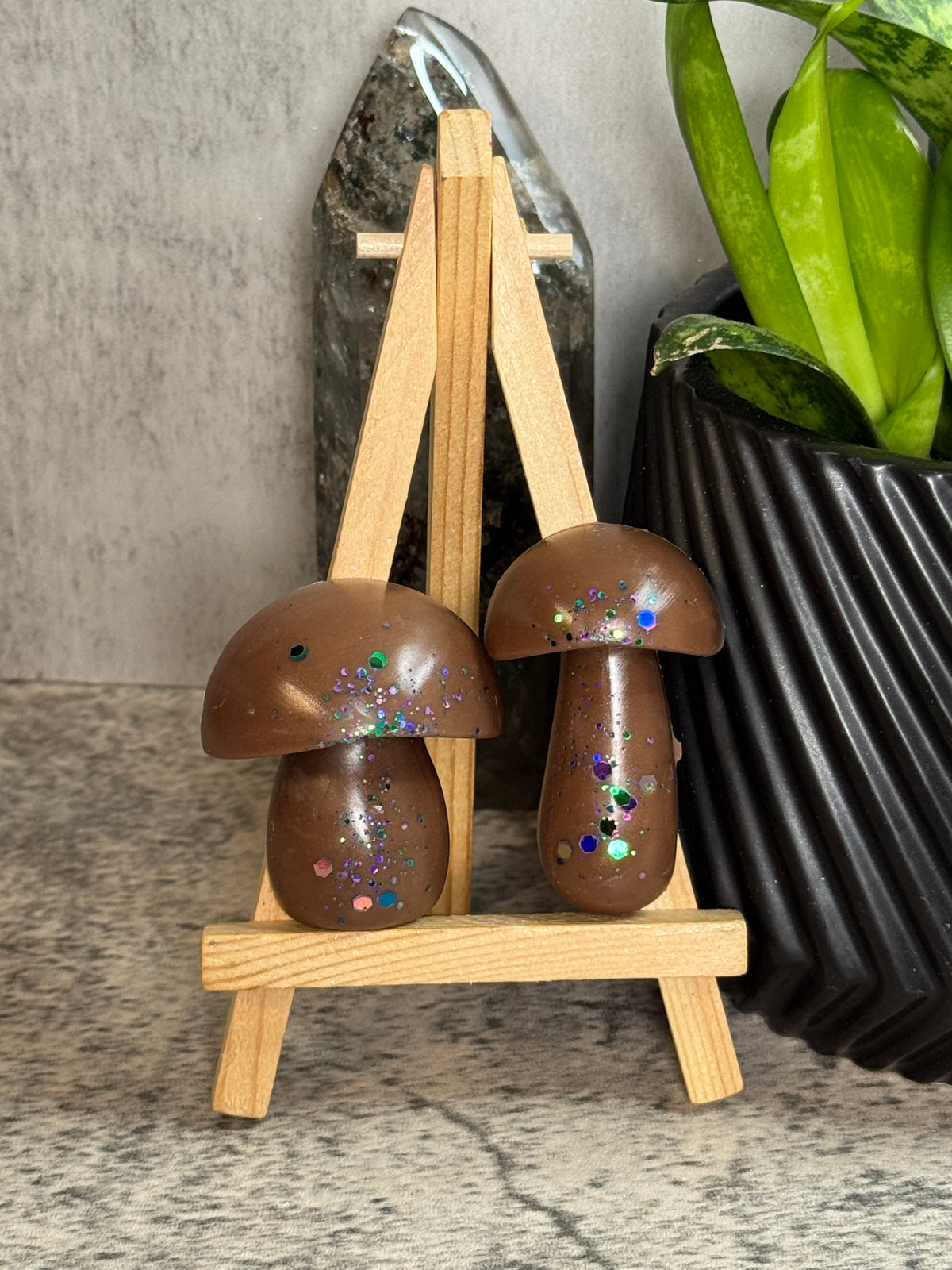 Two brown mushroom-shaped decorative objects with embedded glitter, displayed on a small wooden easel against a gray wall with a plant pot in the background.