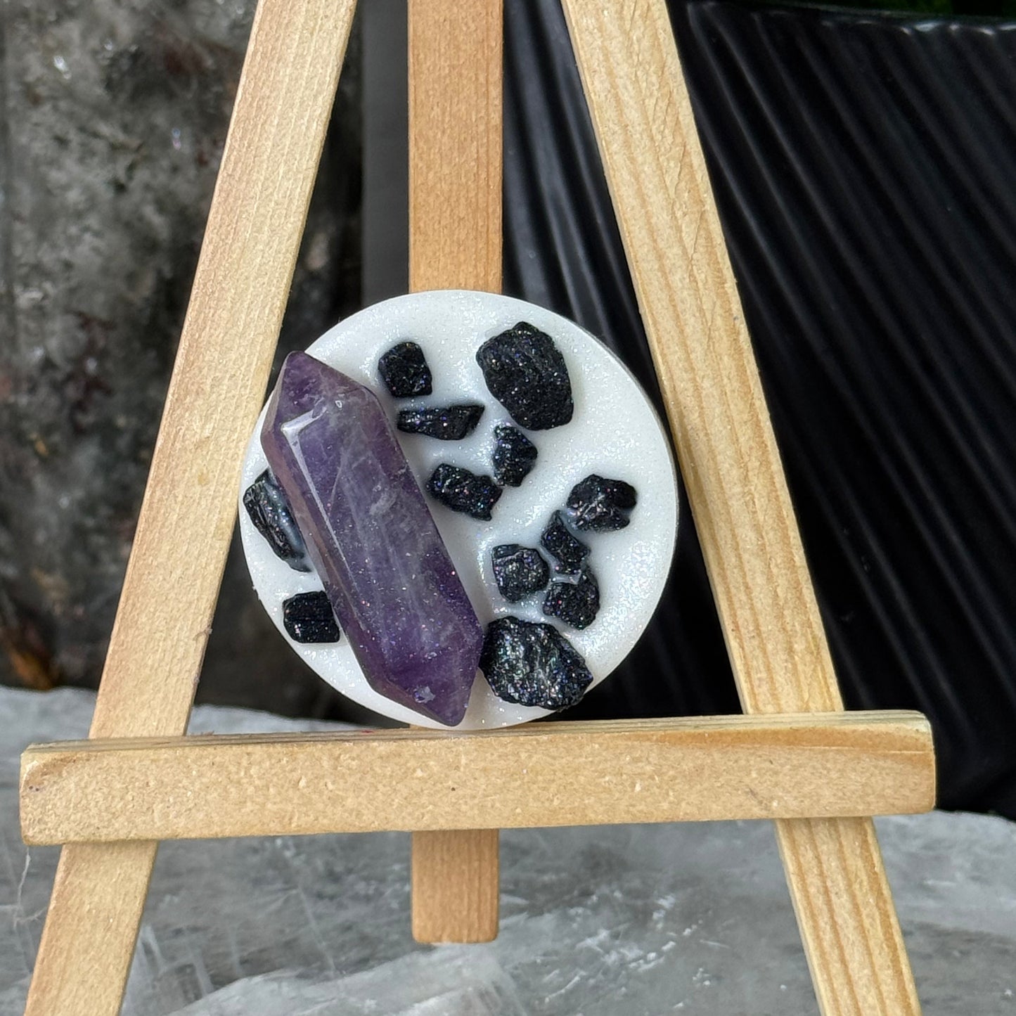 Decorative display with black stones, purple crystal, and white plate on a wooden stand.