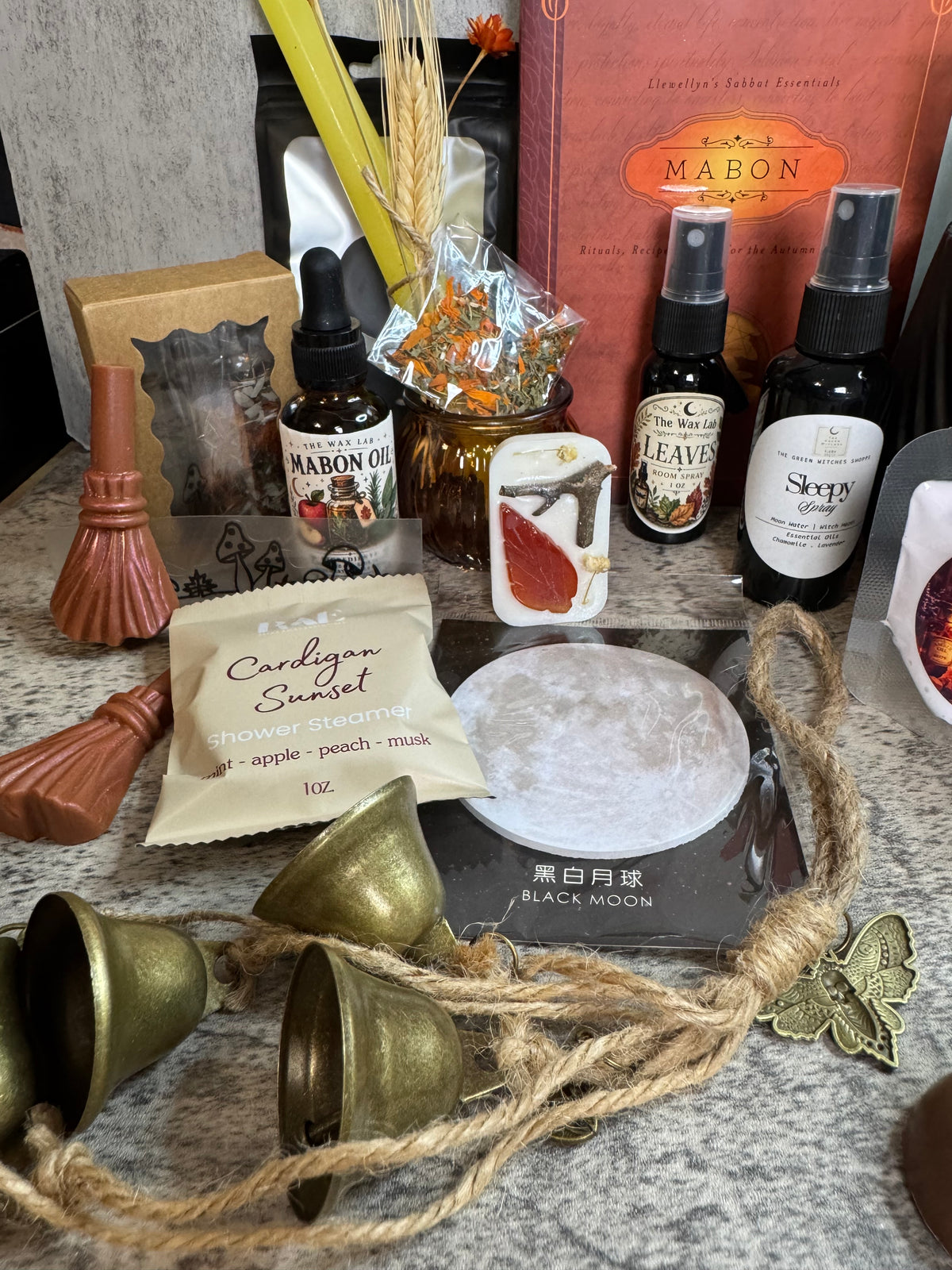 A tabletop arrangement of several wax candle products and related accessories, including a large pink MA BON box, two dark bottles labeled MA BON OIL and SKY'S, a small card labeled LEAVES, a cream packet reading Cardigan Sunset, brass bells, rope, and a white circular wax melt or soap piece on a black tray.