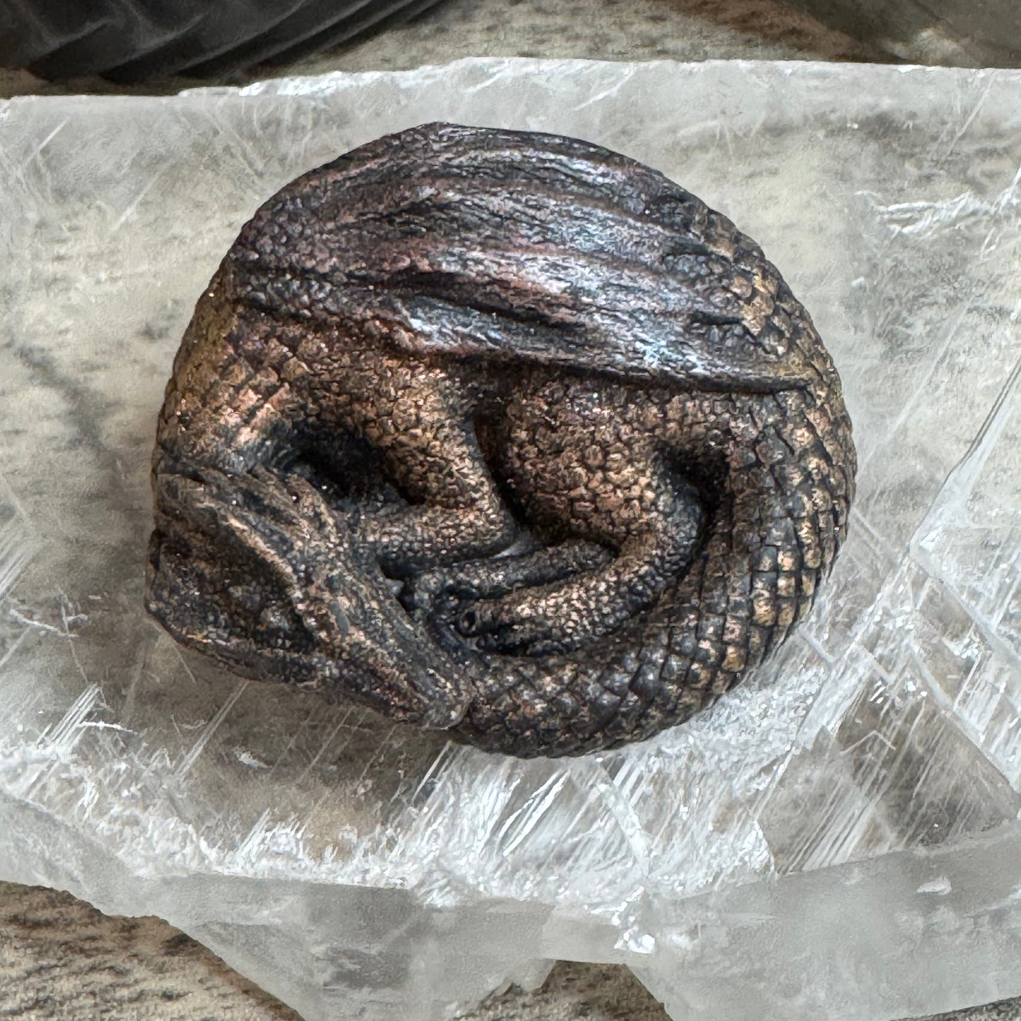 A small sculpted dragon figurine with textured scales in dark metallic bronze and black tones, resting on a clear crystal cluster over a speckled stone surface.