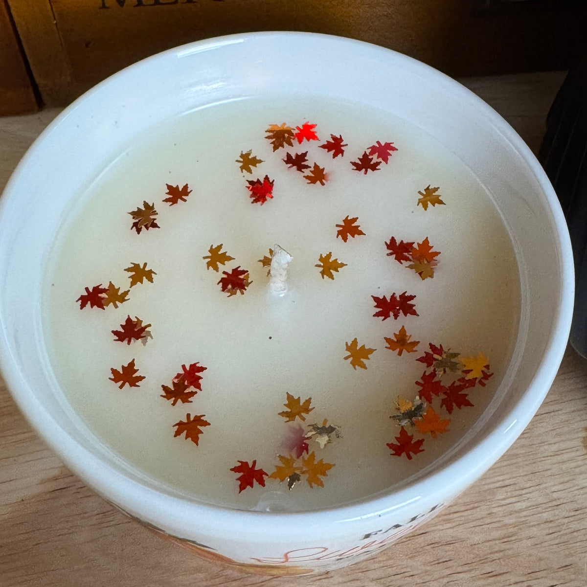 A white ceramic candle bowl filled with creamy off-white wax and decorated with dried red, orange, and yellow maple leaves, set on a light wood surface with a wooden box in the background.