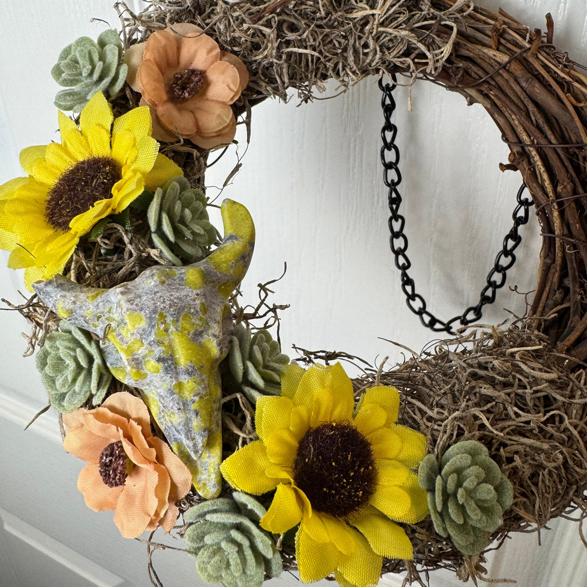 A rustic grapevine wreath decorated with two yellow sunflowers, peach anemone flowers, green succulents, a gray ceramic bird feeder, and brown dried moss, hanging on a white paneled door.