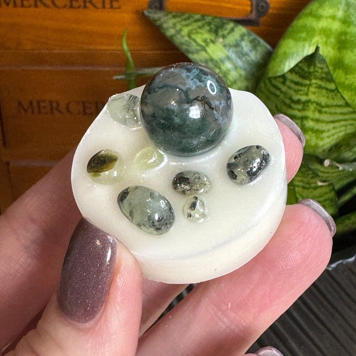 A hand with mauve glitter nail polish holds a white circular stone holder containing a large dark green polished gemstone and several smaller polished stones. In the background, a wooden drawer with the word 'MERCERIE' is visible alongside green snake plants.