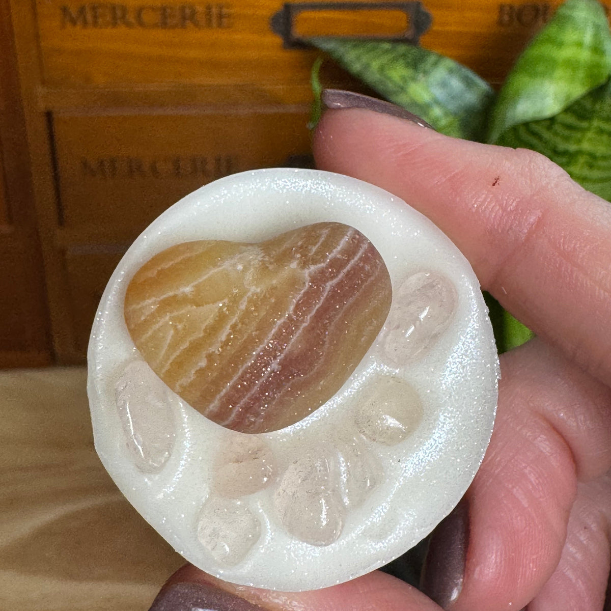 A hand holds a white circular stone jewelry piece featuring a polished heart-shaped stone in the center with a brown and beige striped pattern, surrounded by small clear crystal-like beads. A wooden drawer with the word 'MERCERIE' is blurred in the background.
