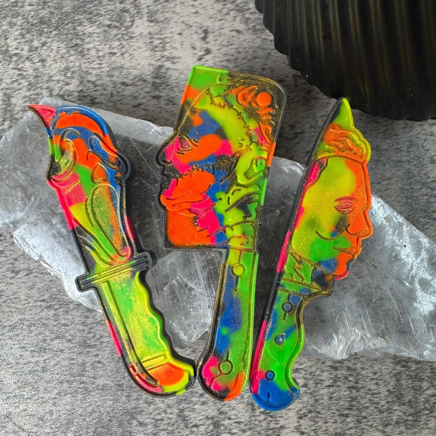 Three colorful novelty knife-shaped items with a neon marbled pattern in green, yellow, orange, blue, and pink, arranged on a clear plastic sheet over a gray textured surface.