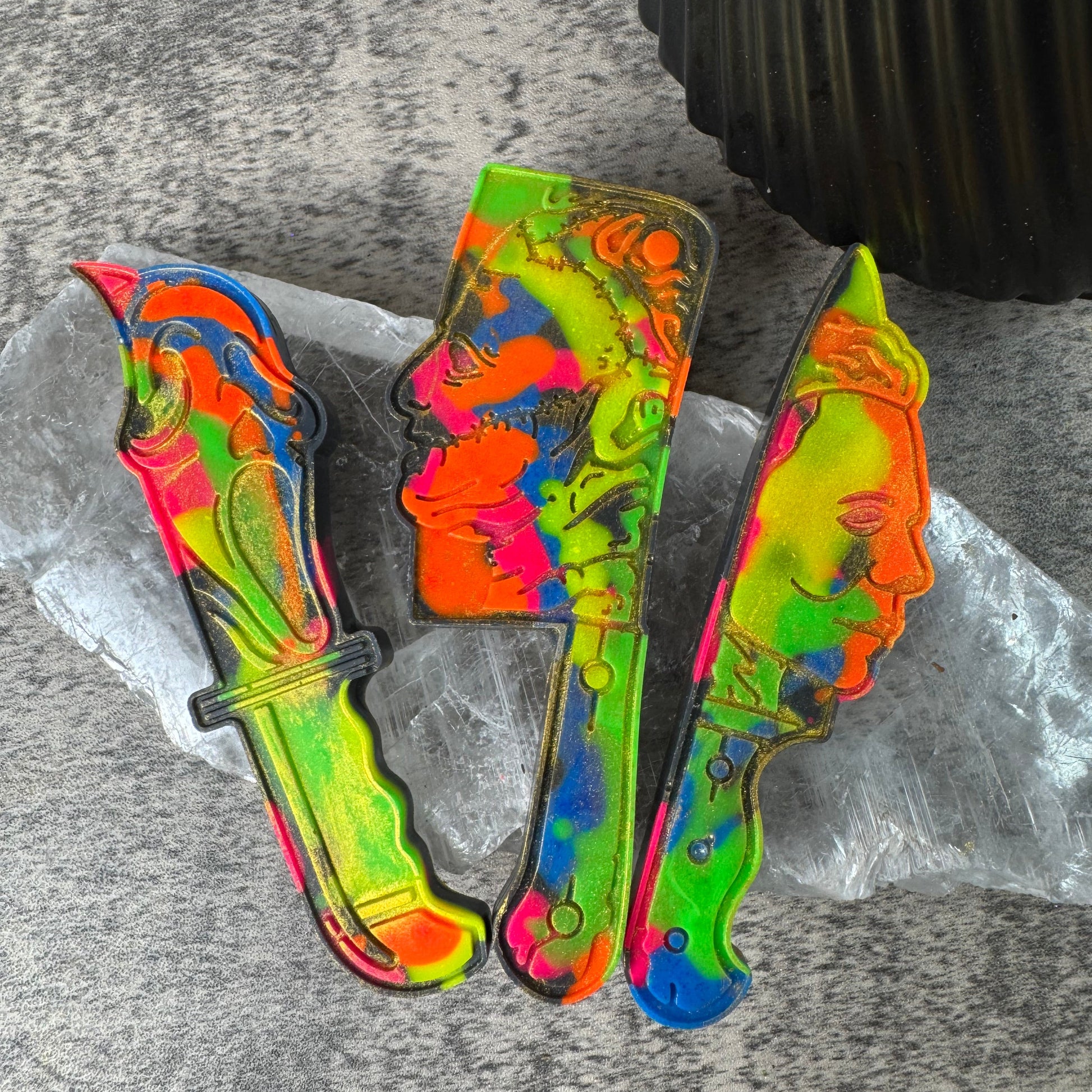 Three colorful novelty knife-shaped items with a neon marbled pattern in green, yellow, orange, blue, and pink, arranged on a clear plastic sheet over a gray textured surface.