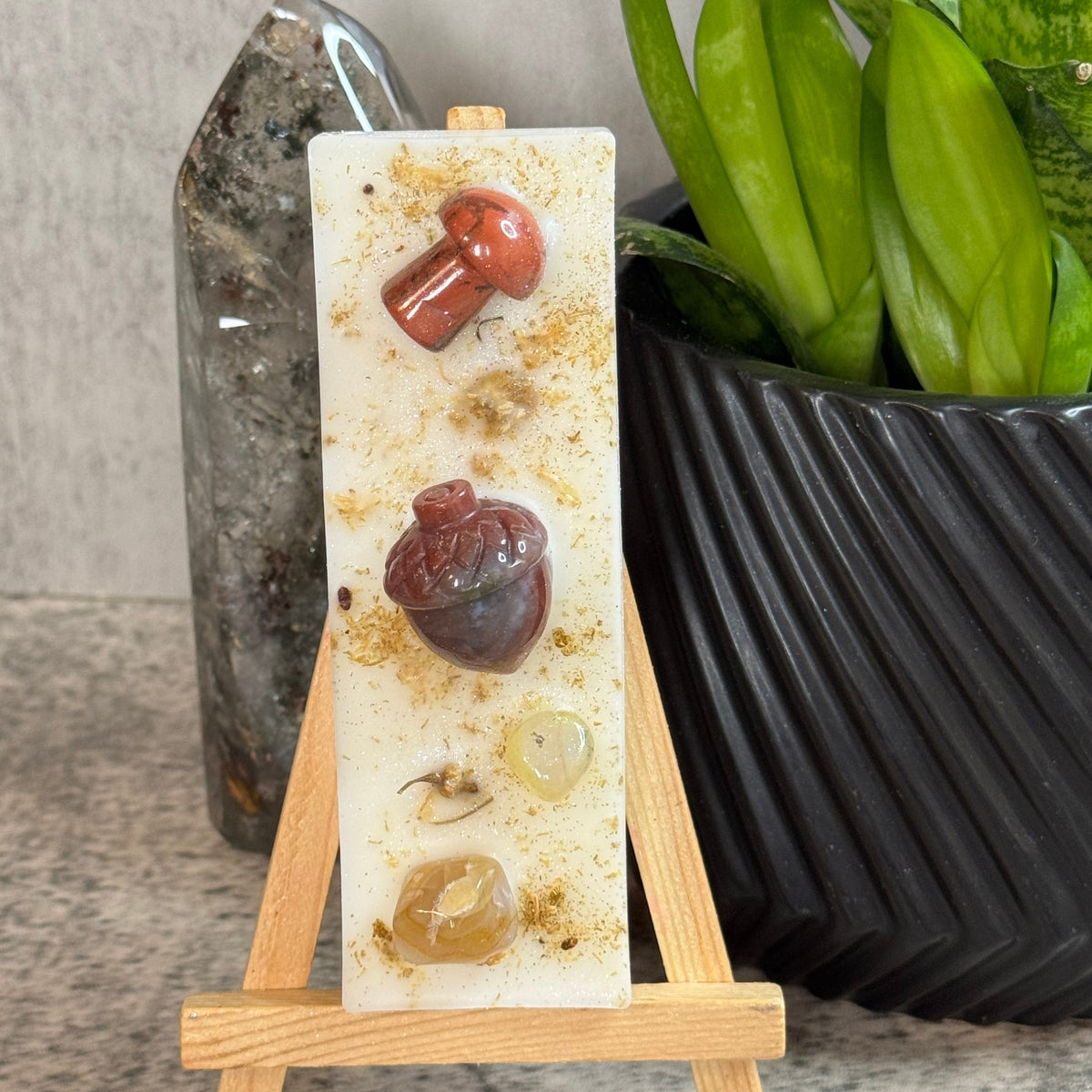 A vertical resin plaque or candle-like decorative piece displayed on a small wooden easel stand. The white resin panel contains embedded natural stones and amber-like inclusions, including reddish-brown and amber pieces.
