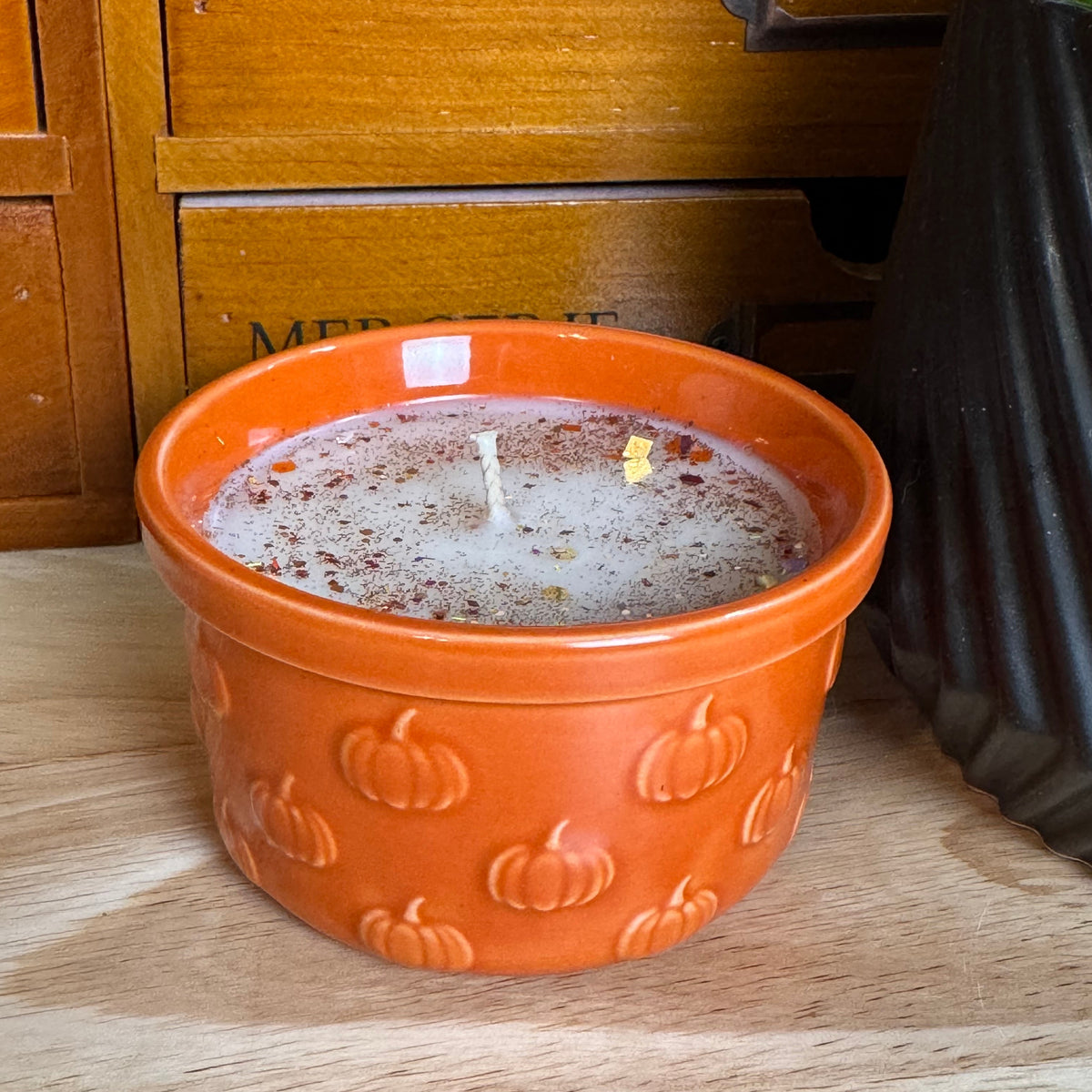 An orange ceramic candle jar with a pumpkin pattern, filled with white wax and a single wick, sitting on a wooden surface in front of a wooden drawer with the word 'MERCERIE' on it.