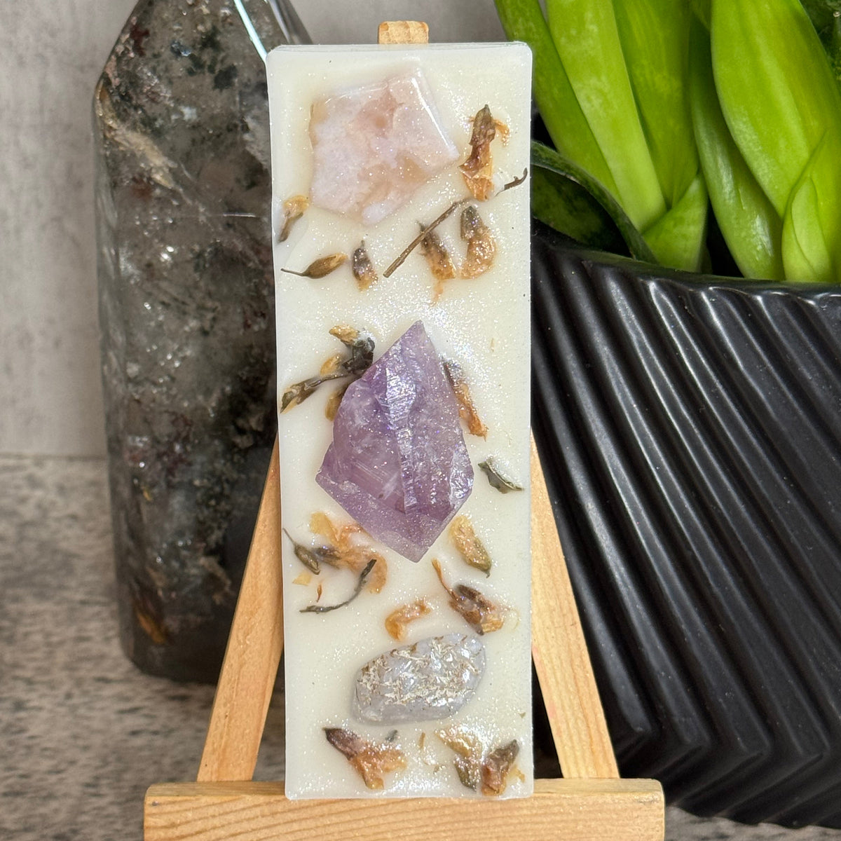 A rectangular white resin candle displayed on a small wooden easel. The candle features embedded natural crystals, including a prominent purple amethyst and smaller pink and silver stones, with dried botanical elements.