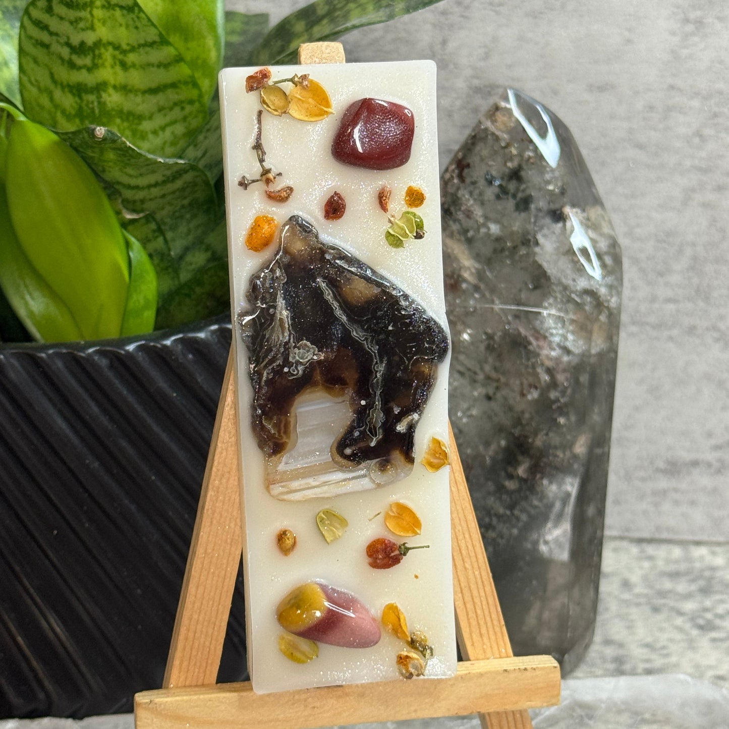 A rectangular cream-colored resin candle displayed on a small wooden easel. The candle features a large dark brown and black animal figure in the center, surrounded by small dried flowers and stones, with a clear crystal embedded near the bottom.
