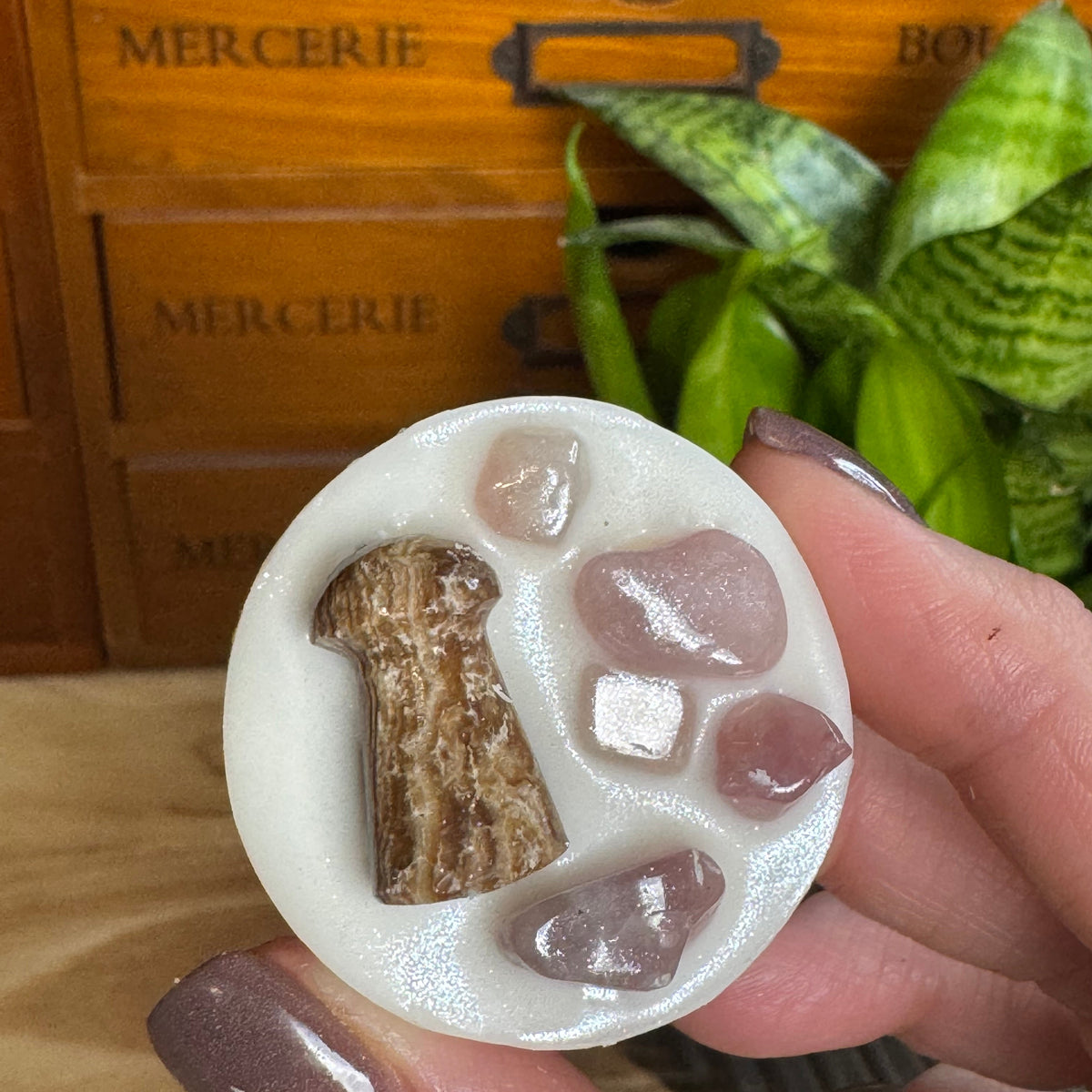 A hand with mauve-painted nails holds a round white resin coaster featuring a brown keyhole and pink cat-eye crystals, with a wooden drawer and green plant in the background.