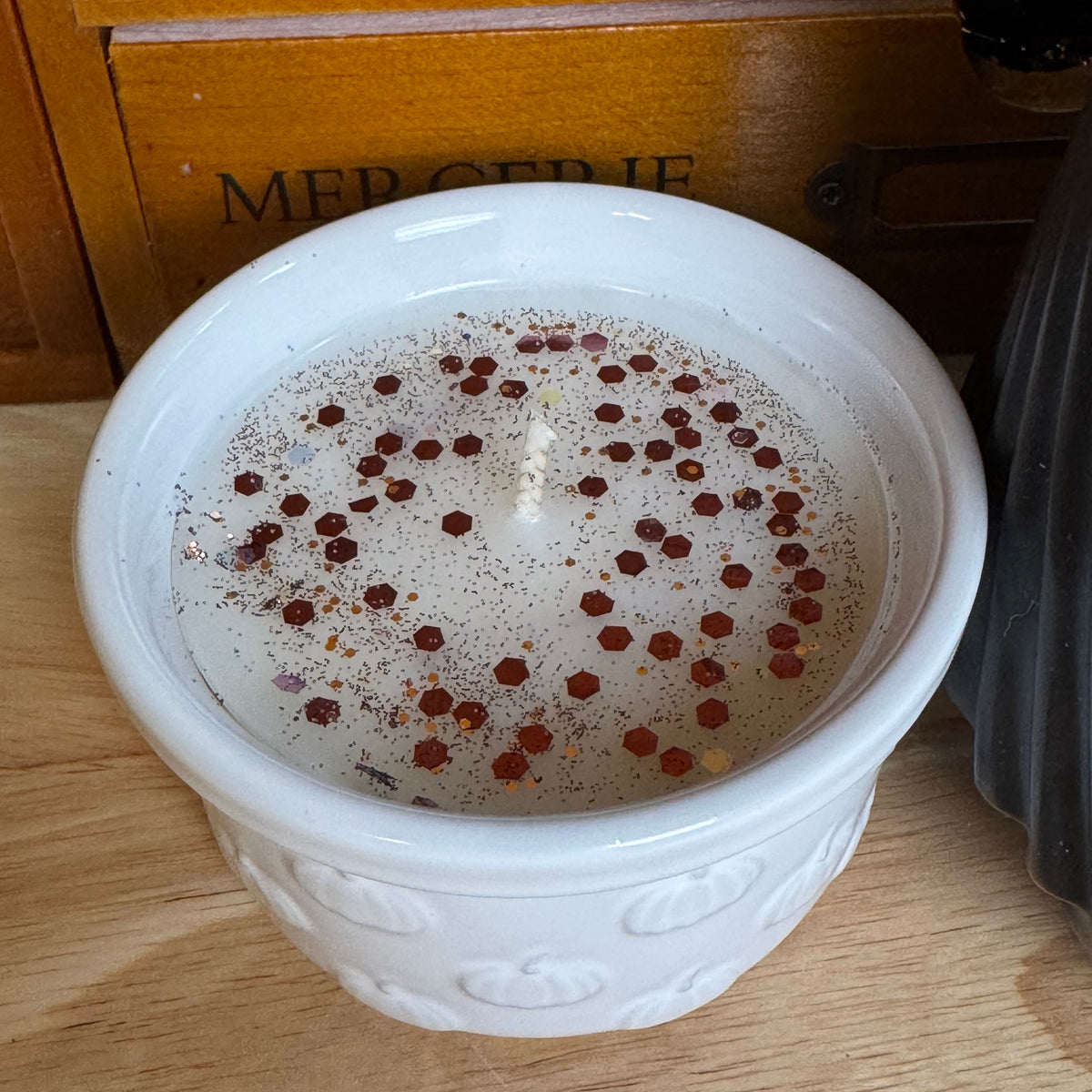 A white ceramic candle jar with a decorative embossed pattern, filled with white wax and topped with scattered red chili flakes, set on a wooden surface with a wooden drawer in the background.