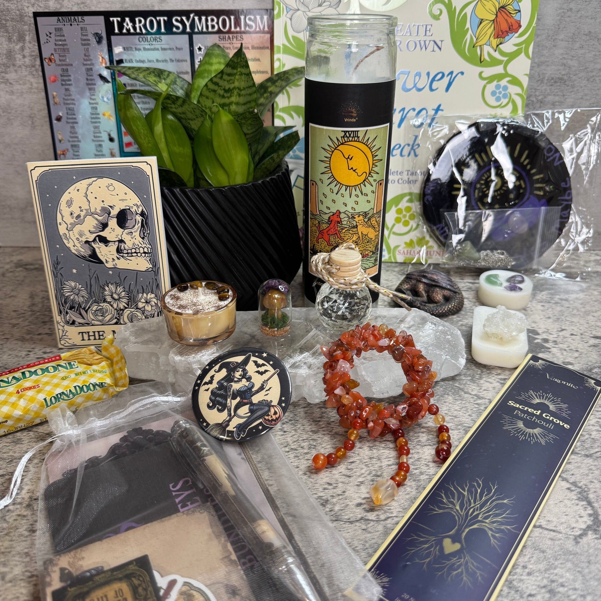 A curated tarot-themed gift set arranged on a gray stone surface, including a tarot card, tarot symbolism card, tarot candle, tarot book, crystals, a red-orange beaded bracelet, a plant in a black basket, and packaged accessories.