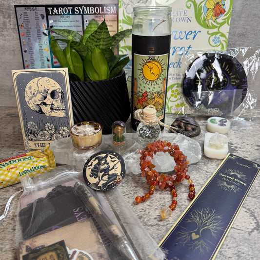 A curated tarot-themed gift set arranged on a gray stone surface, including a tarot card, tarot symbolism card, tarot candle, tarot book, crystals, a red-orange beaded bracelet, a plant in a black basket, and packaged accessories.