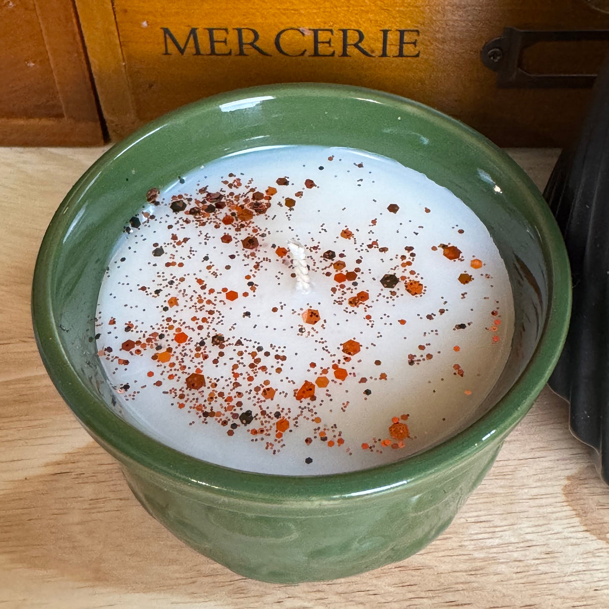 A green ceramic candle holder filled with white wax and topped with orange and black spice flakes, set on a wooden surface with a wooden drawer in the background labeled 'MERCERIE'.