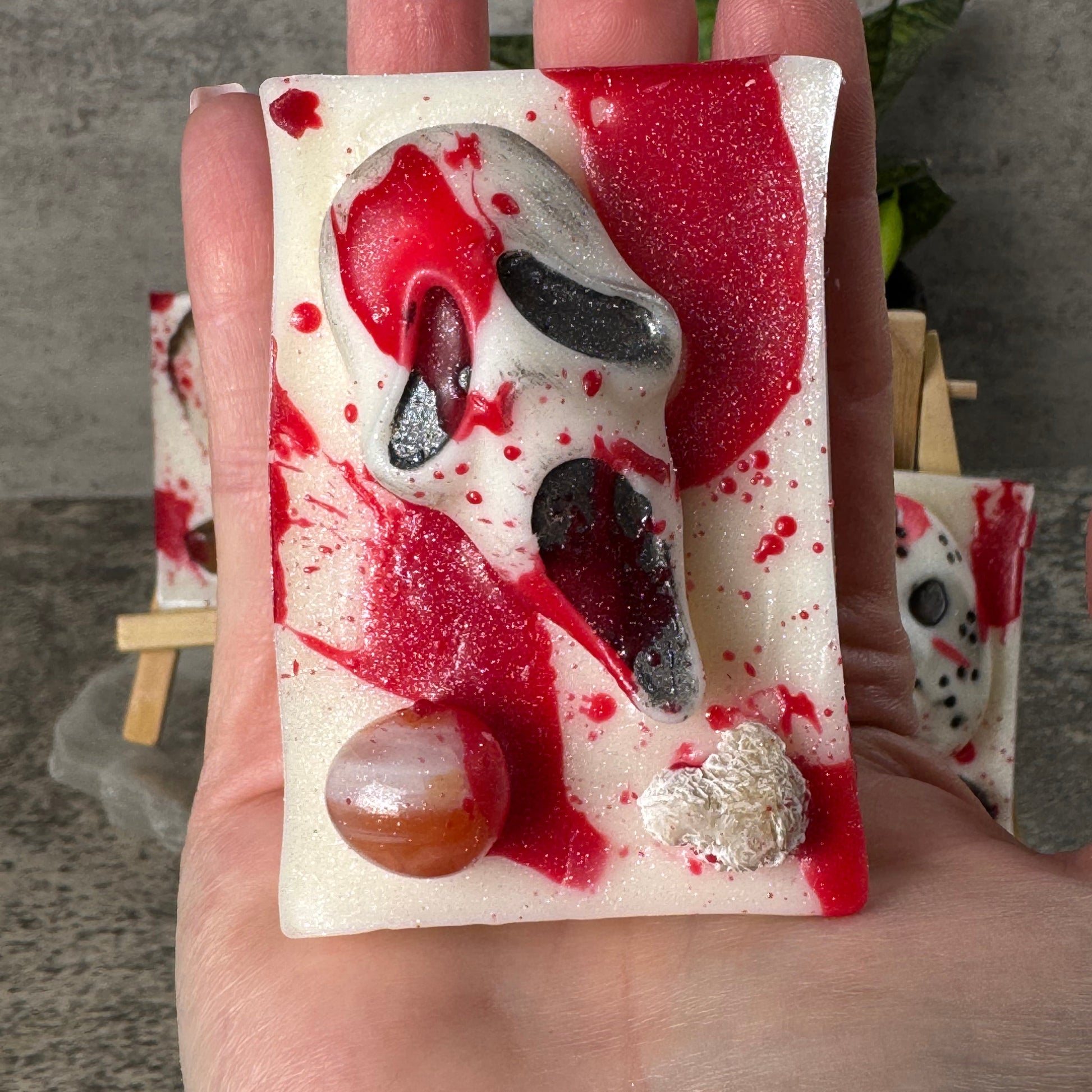 A hand holding a rectangular handmade soap bar featuring a white base with red splatter details, a black and white mask design, and decorative stones.