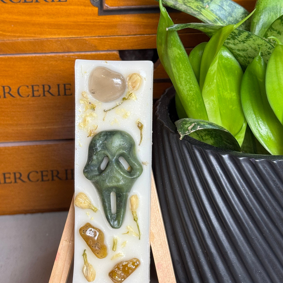 A rectangular white soap bar with a green ghost face and embedded stones, displayed on a small wooden easel. The background features a wooden box with 'CERIE' text and a green potted plant.