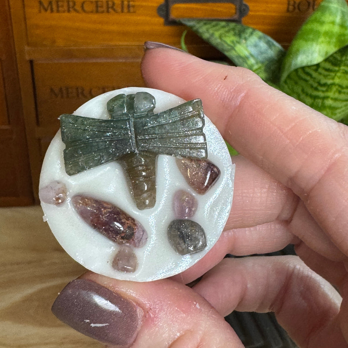 A hand holds a round white resin plaque featuring a green crystal dragonfly and small polished gemstones, with a wooden drawer and green plant leaves in the background.