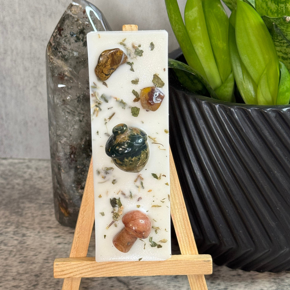 A rectangular white resin plaque or candle holder with embedded natural stones, dried herbs, and a small mushroom, displayed on a small wooden easel. The setup is styled on a stone surface with a crystal cluster and a green potted plant in the background.