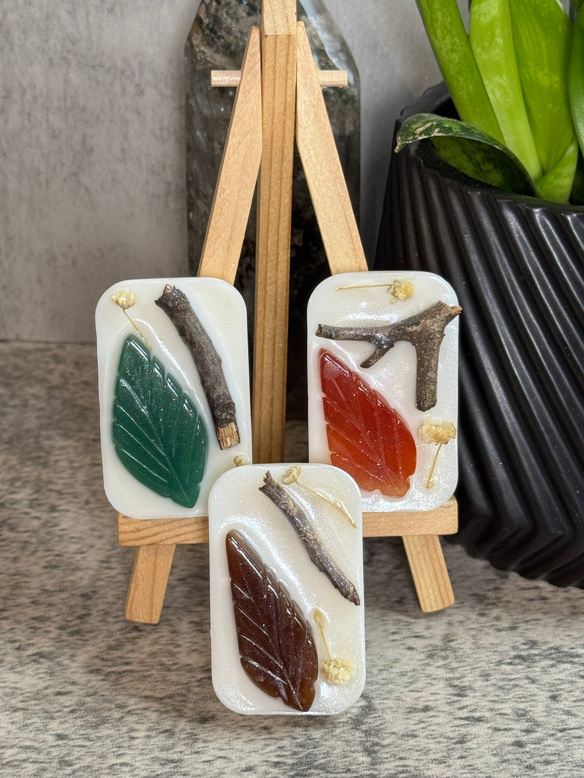 Three rectangular resin keepsakes with dried botanical inclusions, displayed on small wooden easels against a neutral stone background with a potted plant.