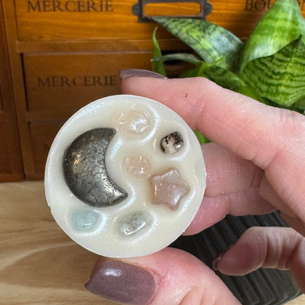 A hand with mauve nail polish holds a round, off-white resin candle featuring a metallic silver crescent moon and small clear and brown gemstone details. The background shows a wooden drawer with 'MERCERIE' text and a green plant.