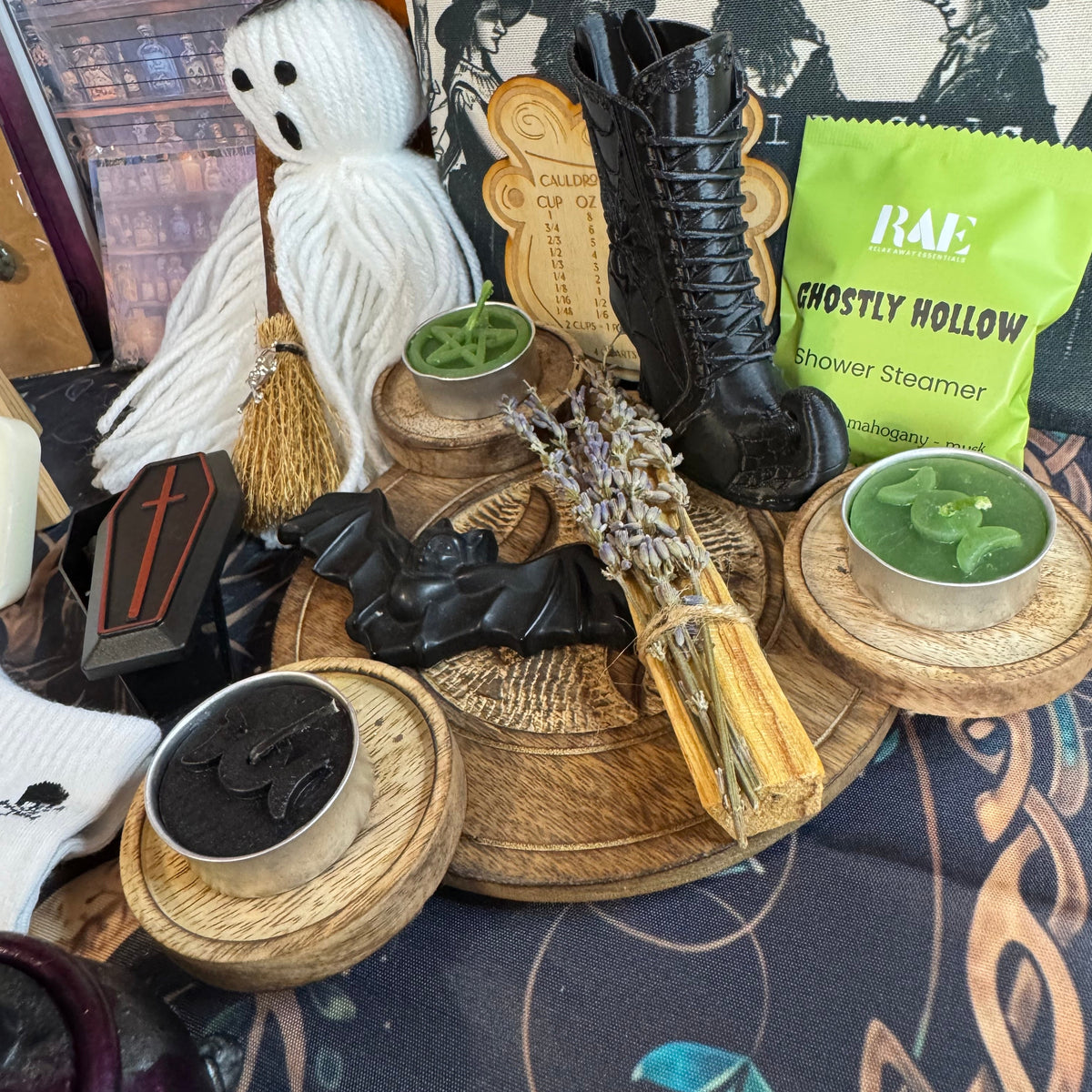 A styled Halloween-themed tabletop display featuring a white yarn ghost doll, black lace-up boots, a witch hat print pillow, a green bag of RAE Ghostly Hollow shower steamers, candles, a broom, and wooden trays on a patterned tablecloth.