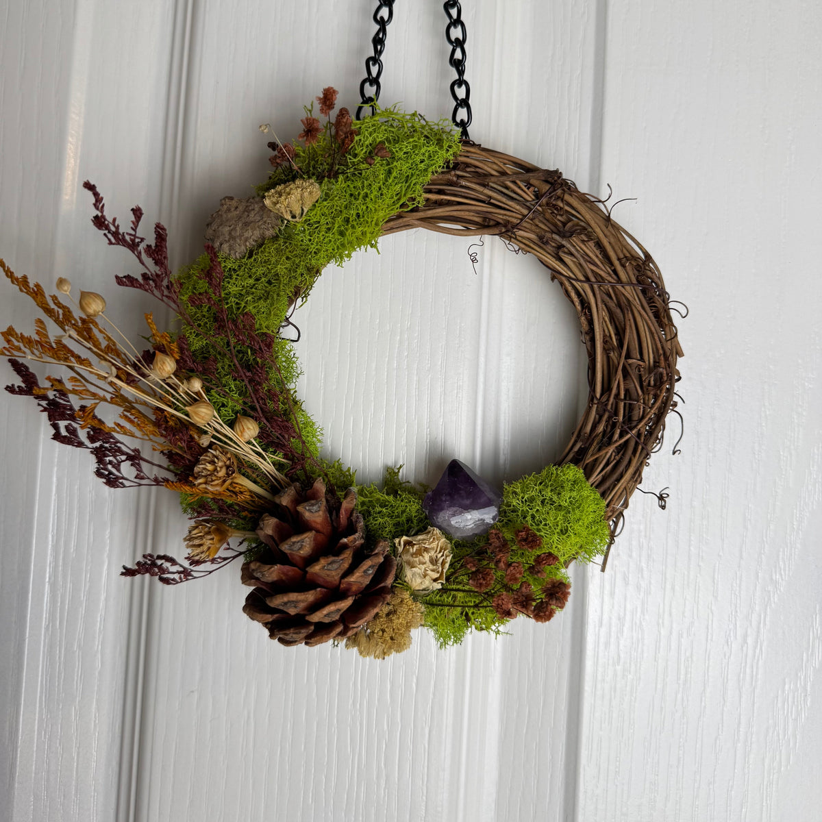 A rustic dried flower wreath hanging on a white paneled door. The wreath is made of natural brown vine and decorated with bright green moss, a large brown pinecone, dried brown and beige flowers, and a small purple crystal.