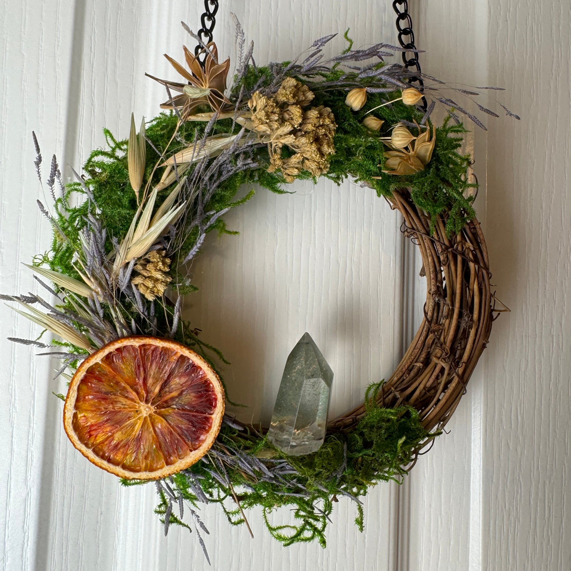 Decorative wreath with greenery, dried fruit, and a crystal on a white door.