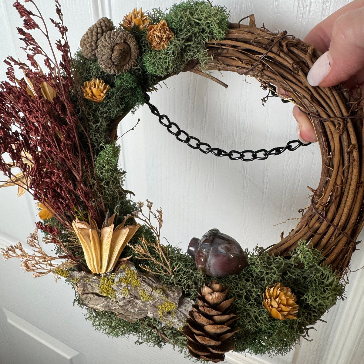 An autumn-themed grapevine wreath decorated with green moss, pinecones, acorns, dried burgundy grasses, and gold-toned leaves, held by a hand against a white paneled door.