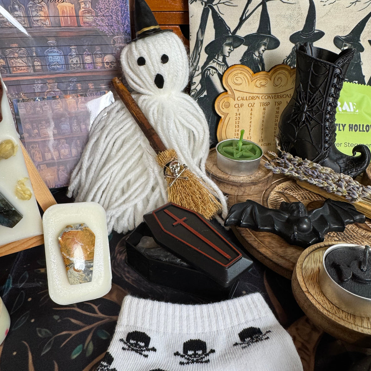 A styled flat-lay of Halloween-themed items, featuring a pair of white crew socks with a black skull-and-crossbones pattern in the foreground, a white fuzzy ghost decoration holding a broom, a small black coffin, a witch hat, and other spooky accessories.
