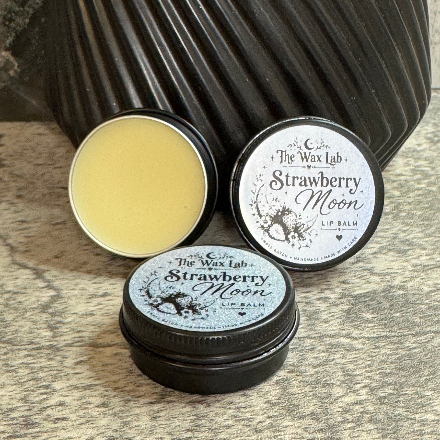 Three round metal tins of The Wax Lab Strawberry Moon lip balm arranged on a textured stone surface with a dark ribbed vase behind them. One tin is open, showing pale yellow balm, while two tins are closed with black and silver lids featuring white text and illustration.