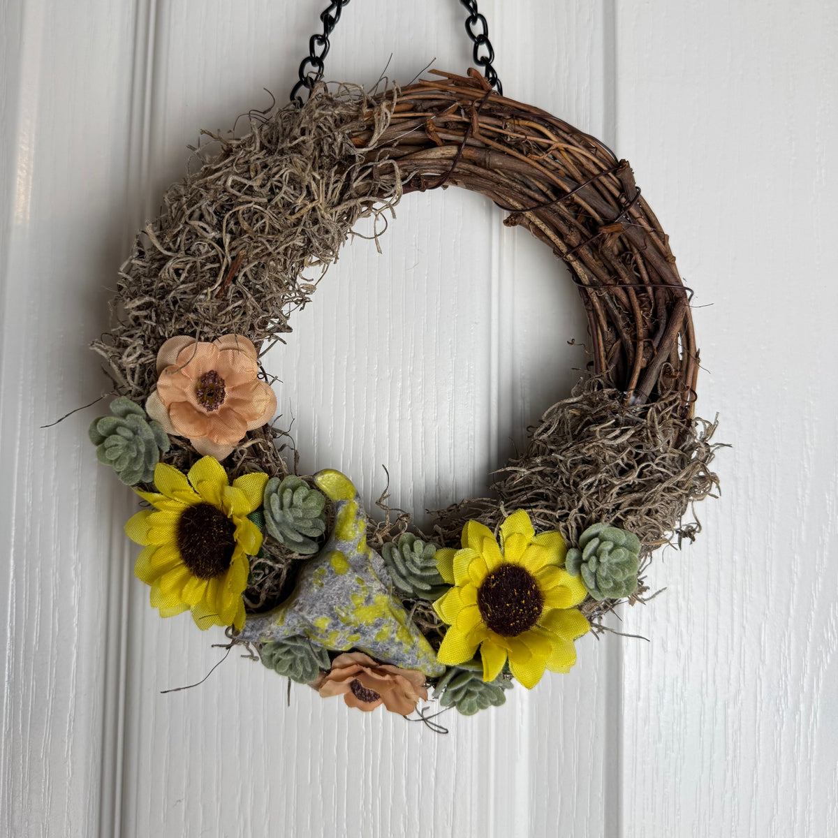 A rustic grapevine wreath hanging on a white paneled door, decorated with faux yellow sunflowers, green succulents, tan dried flowers, and a small gray bird figurine.